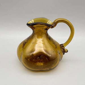 May include: A golden-amber glass pitcher with a rounded body and a curved handle. The pitcher has a small spout and is decorated with tiny air bubbles throughout the glass. The handle is attached near the top and bottom of the pitcher.