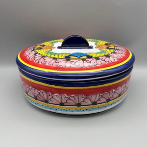May include: A round ceramic serving dish with a lid. The dish is painted with a vibrant floral design in red, yellow, blue, and pink. The lid has a dark blue handle.