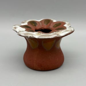 May include: A handcrafted, flower-shaped ceramic vase. The vase features a dark red body with a white and brown floral rim. The interior is black. The vase is approximately 4 inches tall.