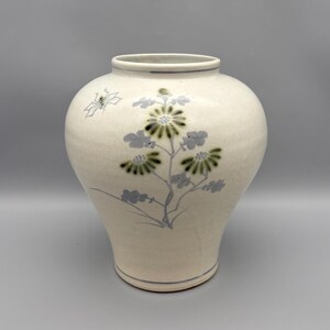 May include: A cream-colored ceramic vase with a rounded body and a narrow neck. The surface features a hand-painted botanical design in shades of green and grey, depicting a flowering plant and a small insect. Thin blue lines circle the rim and base.