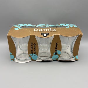 May include: A set of six clear glass teacups in a cardboard box. The box is brown with blue and white floral accents and the word "Damla" printed on it. The teacups are visible through the box's cutouts.