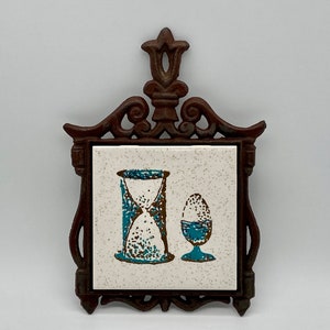 May include: A brown cast iron trivet with an ornate design holds a white ceramic tile with a blue and brown hourglass and egg design.
