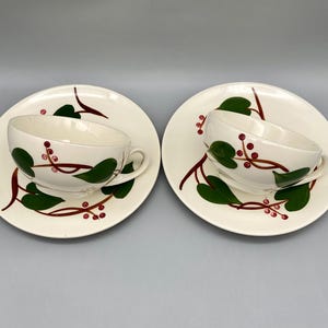 May include: Two white teacups and saucers with a green vine and red berry pattern. The cups are slightly angled and the saucers are facing the camera.