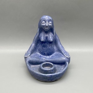 May include: A blue ceramic sculpture of a seated female figure in a meditative pose. The figure has long hair and is seated with legs crossed. A small circular holder is in front of the figure.