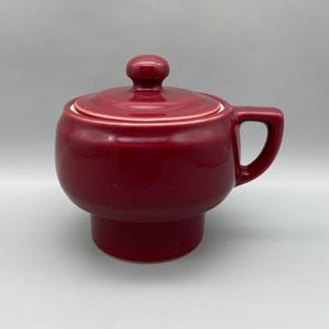 May include: A maroon ceramic sugar bowl with a lid and handle. The bowl has a rounded shape with a pedestal base. The lid has a small knob for easy removal. The handle is a simple, curved design.