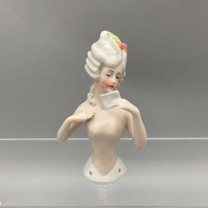 May include: A porcelain doll bust with a white wig and a painted face. The doll is holding a white envelope in her hands. The doll is wearing a white dress and has a floral design on her head.