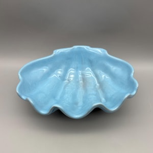 May include: A light blue ceramic seashell-shaped bowl. The bowl has a wavy, scalloped edge and a textured surface. The interior of the bowl is a lighter shade of blue.