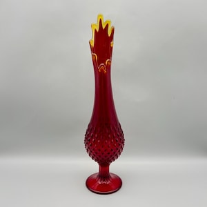 May include: A tall, ruby red glass vase with a textured, hobnail-like pattern on the bulbous lower portion. The vase has a flared, jagged top with a yellow rim. The base is round and the overall shape is elongated.
