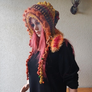 Peut inclure: Un capuchon crocheté à la main, aux tons orange, marron et jaune, avec une bordure en fausse fourrure. Le capuchon présente des accents de perles et un motif texturé, créant un accessoire unique et confortable.