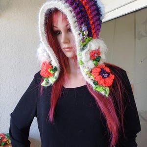 Könnte beinhalten: Eine handgefertigte Häkelhaube in leuchtenden Farben. Die Haube hat ein Muster aus Lila, Orange und Weiß mit einem flauschigen weißen Besatz. Sie ist mit gehäkelten Blumen in Rot-, Orange- und Lilatönen mit grünen Blättern verziert. Die Haube ist zum Tragen bestimmt.