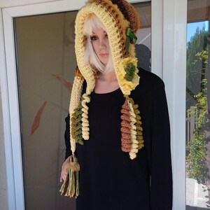 May include: A handmade crochet hood in shades of yellow, brown, and green. The hood features a faux fur trim, a green ribbon, and a decorative flower. The hood has long, dangling tassels with a fur accent.