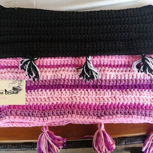 Peut inclure: Une couverture rayée au crochet avec des rayures roses, violettes et blanches. La couverture a une bordure en crochet noir et des glands aux extrémités. La couverture est faite à la main et porte une étiquette qui dit "Anneliese Kreativ".