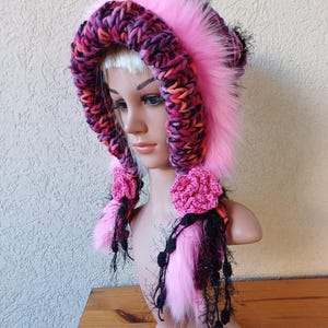 May include: Pink faux fur hooded hat with a multi-coloured crocheted brim and black crocheted tassels. The hat has pink crocheted flowers on each side.