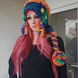 Peut inclure: Un chapeau crocheté coloré avec une longue queue moelleuse. Le chapeau présente un arc-en-ciel de couleurs, notamment l'orange, le vert, le violet et le bleu. La queue est ornée d'un pompon et d'une série de fleurs crochetées.
