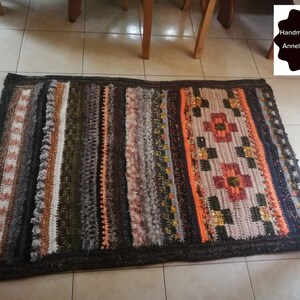 May include: A multi-colored crocheted rug with stripes of brown, orange, green, and gray. The rug has a geometric pattern in the center with a flower design. The rug is handmade and designed by AnnelieseNicoara.