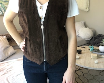 Puede incluir: Un chaleco de ante marrón oscuro con cierre de cremallera plateada. El chaleco tiene cuello en V y dos bolsillos delanteros. La modelo lleva una camiseta blanca y vaqueros azul oscuro.