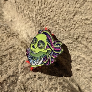 May include: Green and purple enamel pin featuring a skull with a detailed design. The skull has a green face with black eyes and a purple and pink crown. The pin is on a textured beige surface.