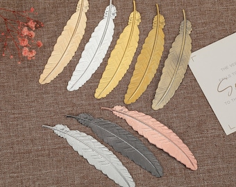 May include: Set of ten gold, silver, and rose gold feather-shaped bookmarks. The bookmarks are all the same size and shape, but they are different colors. They are arranged in a row on a brown fabric background.