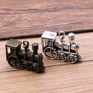 Puede incluir: Dos encantos de locomotoras de tren en miniatura, uno plateado y otro bronce, con ruedas y chimeneas detalladas.