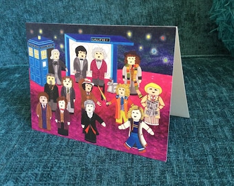 Puede incluir: Tarjeta de felicitación plegada con una colorida ilustración de personajes de dibujos animados, incluyendo una cabina telefónica azul y un letrero que dice "Gallifrey". El fondo muestra una noche estrellada. Los personajes visten varios atuendos.