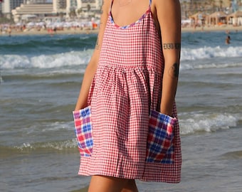 May include: A woman wearing a red and white gingham dress with blue and white plaid pockets stands in the shallow water of a beach. She is wearing sunglasses and has short blonde hair.