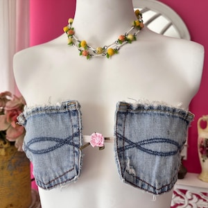 May include: A strapless top constructed from two light blue denim pockets with frayed edges, connected at the center by a thin strap featuring a small pink flower. A necklace with yellow and orange floral beads and green leaves is worn above.