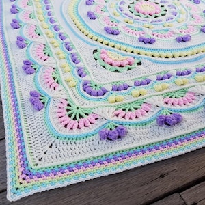 May include: A handcrafted, square crochet blanket in pastel shades. The blanket has a central circular design with scalloped edges, decorated with purple, pink, and yellow floral details. The border features a striped pattern in blue, purple, and yellow.