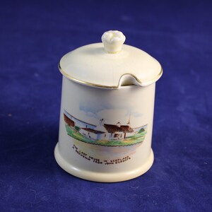 May include: A white ceramic jar with a lid and a gold trim. The jar has a painted scene of a cottage with the text "The Old House in Scotland, Souvenir from your holidays".