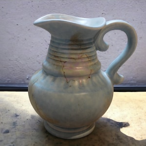 May include: A light blue ceramic pitcher with a handle and a textured surface. The pitcher has a faded, light pink and brown design.