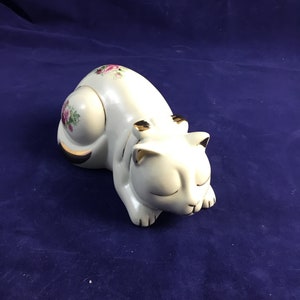 May include: A white ceramic cat figurine with pink and green floral details and gold accents. The cat is curled up in a sleeping position.