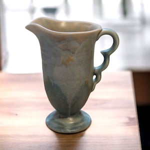 May include: A blue and white ceramic pitcher with a decorative handle. The pitcher has a floral design and a footed base.