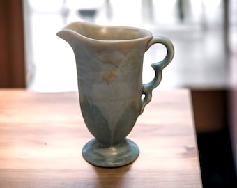 May include: A blue and white ceramic pitcher with a decorative handle. The pitcher has a floral design and a footed base.