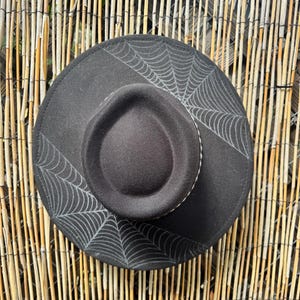 May include: Black fedora hat with a spiderweb design. The hat has a round crown and a wide brim, with a white stitched band around the base of the crown. The spiderweb design is printed on the brim.