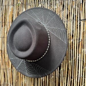 May include: A black fedora hat with a spiderweb design on the brim. The hat has a silver-coloured band around the crown. The hat is set against a natural bamboo fence.