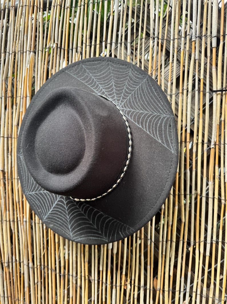 May include: A black fedora hat with a spiderweb design on the brim. The hat has a silver-coloured band around the crown. The hat is set against a natural bamboo fence.