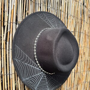 May include: Black fedora hat with a silver beaded band and a spiderweb design. The hat is made of a felt-like material and has a wide brim. The spiderweb design is in a light grey colour.
