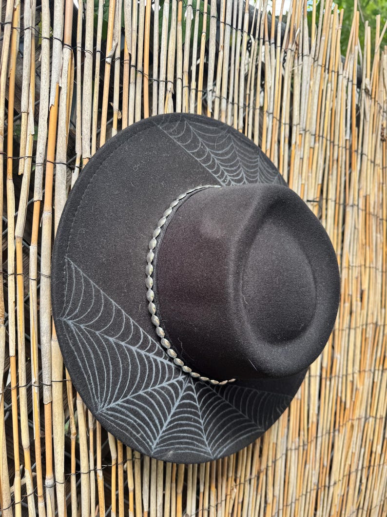 May include: Black fedora hat with a silver beaded band and a spiderweb design. The hat is made of a felt-like material and has a wide brim. The spiderweb design is in a light grey colour.