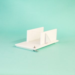 May include: White plastic soap cutter with a clear plastic guide. The cutter has a handle and a sliding mechanism for cutting soap bars into even slices.