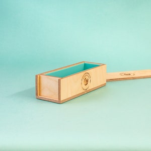 May include: A wooden soap mold with a light blue silicone insert. The mold has a label that reads "The Tradition of Heritage - Handcrafted in USA - Premium Soap Box".