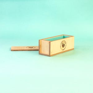 May include: A rectangular wooden soap mold with a light blue interior and a separate lid. The mold has a natural wood finish and a circular logo on the side. The lid is also made of wood.