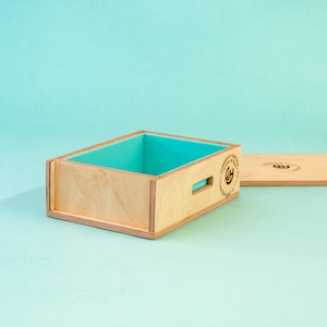 Puede incluir: Una caja de madera con un revestimiento de silicona turquesa y una tapa de madera. La caja tiene un asa cortada en el lateral y un logotipo circular en la tapa que dice "Hecho a mano en los EE. UU., Jabón de primera calidad".