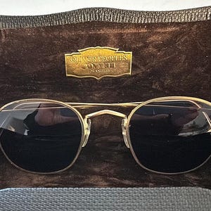 May include: Gold-rimmed sunglasses with black lenses in a brown velvet case. The case has a gold label that reads "Oliver Peoples, Los Angeles".