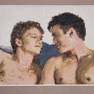 May include: A cross-stitch embroidery piece depicting two individuals facing each other. The figures are rendered in shades of tan, brown, and pink against a dark blue background, showcasing detailed needlework on a light-colored fabric grid.