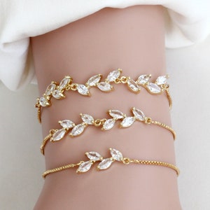 May include: Three gold-toned bracelets with a leaf design, each adorned with clear, sparkling crystals. The bracelets are delicate and elegant, perfect for adding a touch of glamour to any outfit. The bracelets are shown on a wrist.