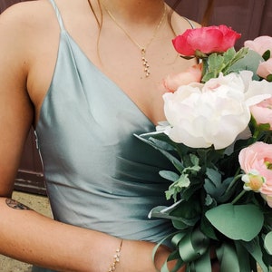 May include: A sage green satin slip dress with thin spaghetti straps and a draped bodice, paired with a gold chain necklace featuring a leaf-shaped pendant and a matching gold leaf bracelet, held alongside a bouquet of white and pink flowers.