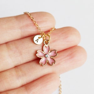 May include: A gold chain necklace with a pink cherry blossom charm and a small gold disc charm with the letter 'A' engraved on it.
