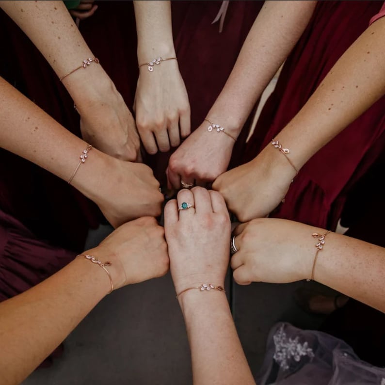 May include: A collection of delicate rose gold bracelets, each featuring a small cluster of clear, leaf-shaped stones. The bracelets are worn on multiple wrists arranged in a circle, with one wrist also displaying a silver ring with a teal stone.