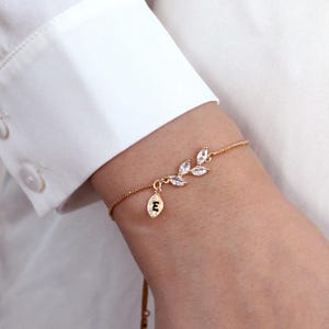 May include: A gold-toned chain bracelet featuring a delicate branch motif adorned with four clear marquise-cut stones and a small, dangling leaf-shaped charm engraved with the uppercase letter E, secured with a thin, adjustable metal chain.