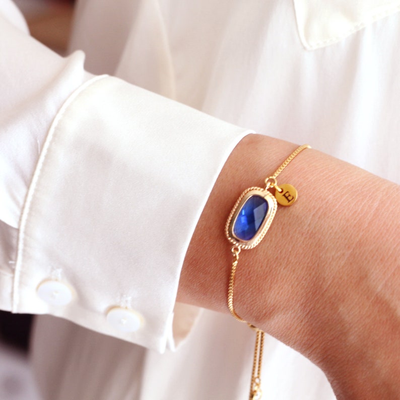 May include: A gold chain bracelet with a blue gemstone and a gold charm with the letter "E" on it.