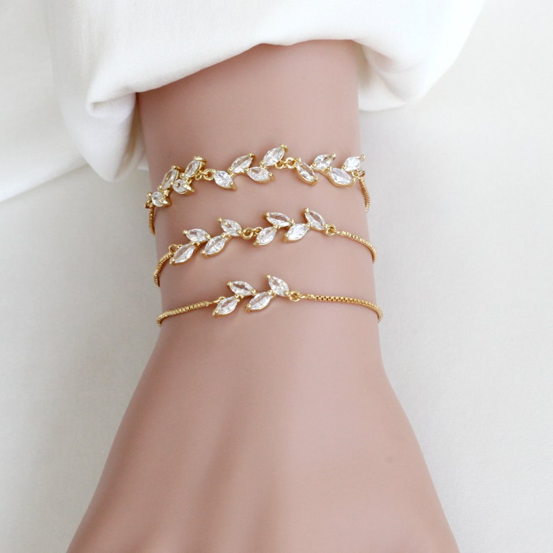 May include: Three gold-toned chain bracelets featuring marquise-cut clear cubic zirconia stones arranged in leaf-like patterns. Each bracelet has a delicate box chain design with adjustable sliders, showcasing a shimmering metallic finish and clear stones.